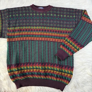 May include: A long-sleeve crew neck sweater featuring a multi-colored geometric pattern in shades of burgundy, forest green, and mustard yellow. The knit garment displays horizontal bands of repeating triangles, diamonds, and vertical stripes.