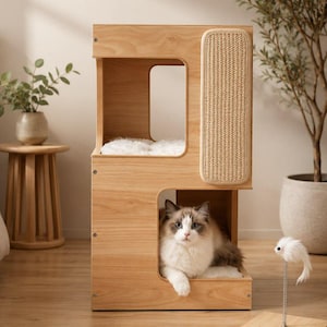 May include: A light wood cat tower featuring two levels, a vertical sisal scratching panel on the side, and a fluffy white cushion on the top platform. A long-haired cat with white and brown fur is resting inside the lower open compartment.