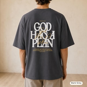 May include: A dark grey short-sleeve t-shirt featuring white block text on the back that reads "GOD HAS A PLAN" with gold cursive script overlaying the text. Below the main graphic, smaller gold text displays "JEREMIAH 29:11" and a Bible verse.