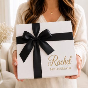 May include: A square white gift box featuring a wide black satin ribbon tied in a large bow. The box is printed with the name Rachel in a gold cursive script font, followed by the word BRIDESMAID in a simple gold sans-serif capital letter font.