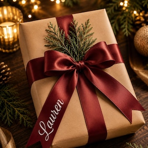 May include: A rectangular gift box wrapped in plain brown paper, tied with a wide, deep burgundy satin ribbon featuring a large bow. The name Lauren is printed in white script on one ribbon tail, accented by a small sprig of green cedar foliage.