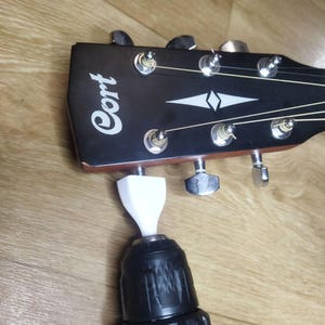 May include: A black Cort guitar headstock with silver tuning pegs and strings. A white adapter is attached to a black drill, positioned to turn the tuning pegs. The guitar headstock has a white diamond design.