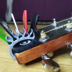 May include: Close-up of a guitar headstock with a variety of colorful guitar picks held in a gray pick holder. The guitar headstock is dark brown with silver tuning pegs and strings. The pick holder has a stylized eye design.