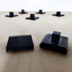 May include: A collection of small, black plastic components are displayed on a light-coloured wooden surface. The components include square bases with a central post and a rectangular piece with many small, raised dots.