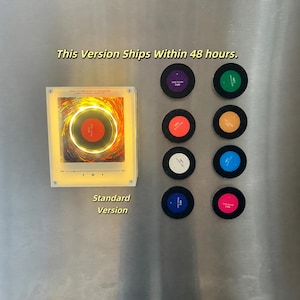 May include: A square, illuminated acrylic display featuring a central circular graphic, accompanied by eight small, circular, multi-colored NFC discs arranged in two vertical columns. Text on the display reads This Is The Darrell Domain Of Ordinary Life.
