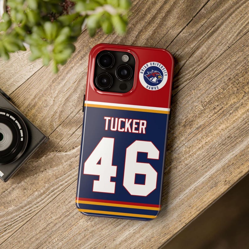 May include: A red, white, and blue phone case featuring the name "Tucker" and the number "46" in white. The case also displays the Briar University Hawks logo. The phone case is on a wooden surface.