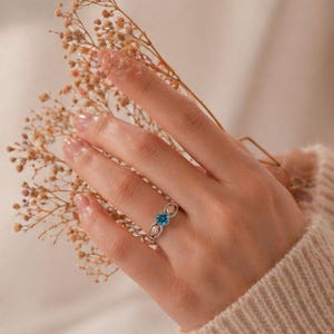 Puede incluir: Un anillo plateado con una piedra preciosa azul redonda en el centro. El anillo tiene un dise&ntilde;o decorativo con detalles en oro rosa. El anillo est&aacute; en la mano de una persona, con flores secas en el fondo.