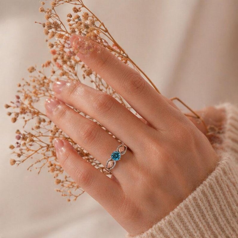 Puede incluir: Un anillo plateado con una piedra preciosa azul redonda en el centro. El anillo tiene un dise&ntilde;o decorativo con detalles en oro rosa. El anillo est&aacute; en la mano de una persona, con flores secas en el fondo.