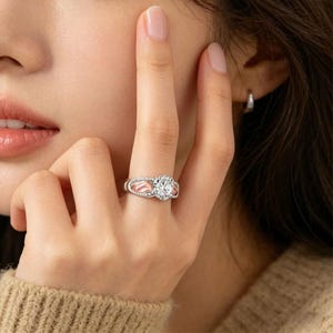 Puede incluir: Un anillo plateado con una piedra preciosa grande, clara y redonda como pieza central. El anillo tiene una banda decorativa con detalles en rosa y piedras claras m&aacute;s peque&ntilde;as. El anillo est&aacute; en un dedo.