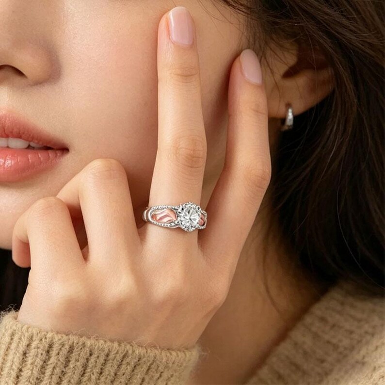 Puede incluir: Un anillo plateado con una piedra preciosa grande, clara y redonda como pieza central. El anillo tiene una banda decorativa con detalles en rosa y piedras claras m&aacute;s peque&ntilde;as. El anillo est&aacute; en un dedo.