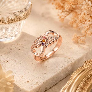 May include: Rose gold ring with an infinity symbol design, featuring wing details and three small gemstones in pink, orange, and purple. The ring is set on a textured surface, showcasing its intricate details and elegant style.