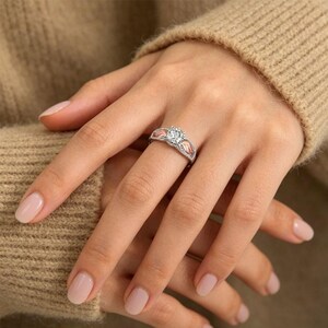 Puede incluir: Un anillo plateado con una gema grande, redonda y transparente en el centro. El anillo tiene un dise&ntilde;o decorativo con detalles en rosa en los laterales. El anillo est&aacute; en un dedo, con la mano apoyada en un su&eacute;ter beige.