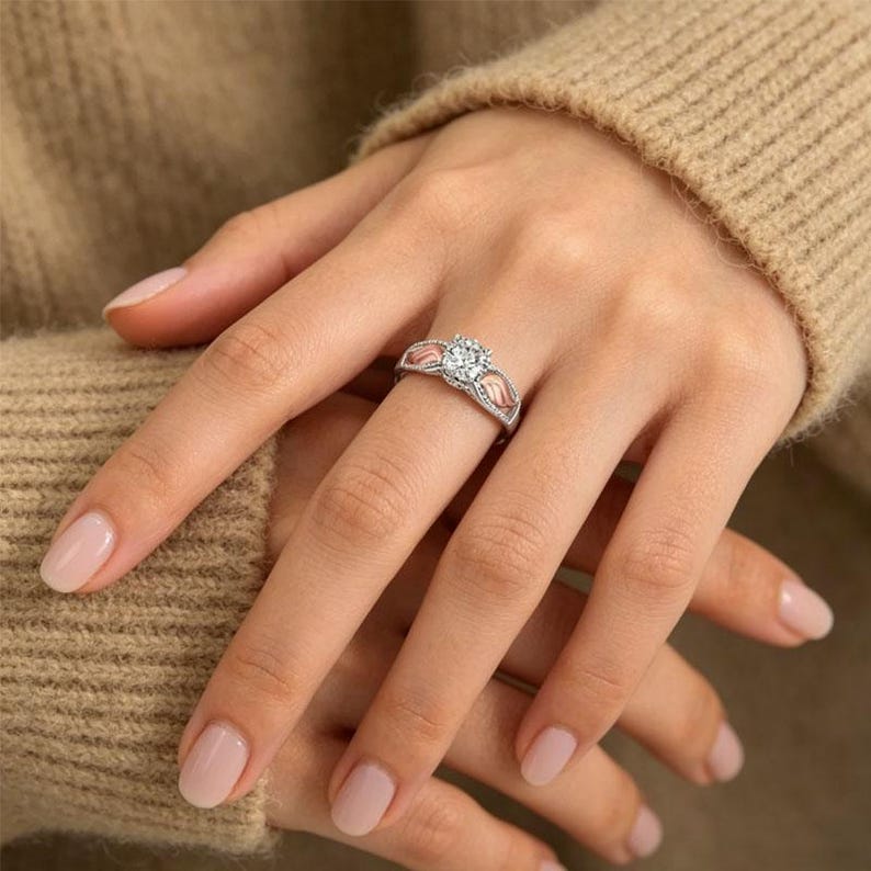 Puede incluir: Un anillo plateado con una gema grande, redonda y transparente en el centro. El anillo tiene un dise&ntilde;o decorativo con detalles en rosa en los laterales. El anillo est&aacute; en un dedo, con la mano apoyada en un su&eacute;ter beige.