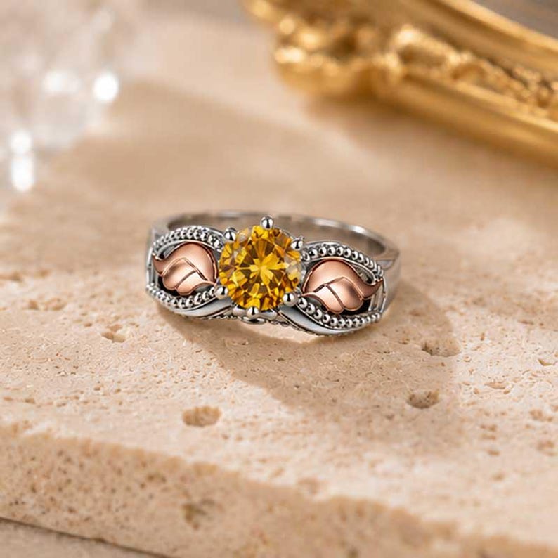 Puede incluir: Un anillo plateado con una gran piedra preciosa amarilla redonda en el centro. El anillo presenta detalles de hojas de color oro rosa a ambos lados de la piedra preciosa. La banda est&aacute; detallada con peque&ntilde;os detalles de cuentas, creando un dise&ntilde;o elegante e inspirado en la naturaleza.