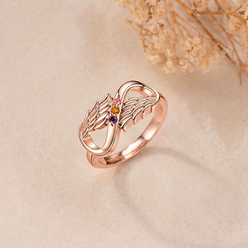 May include: Rose gold ring featuring an infinity symbol design with wing accents. The ring has three small, colorful gemstones. The ring is a jewelry item.