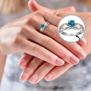 Puede incluir: Un anillo plateado con una piedra preciosa azul redonda en el centro. El anillo tiene un dise&ntilde;o decorativo con detalles en oro rosa. La palabra "Dates" est&aacute; grabada en la banda. El anillo se muestra en un dedo.