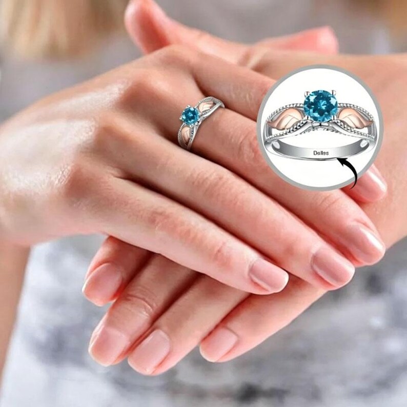 Puede incluir: Un anillo plateado con una piedra preciosa azul redonda en el centro. El anillo tiene un dise&ntilde;o decorativo con detalles en oro rosa. La palabra "Dates" est&aacute; grabada en la banda. El anillo se muestra en un dedo.