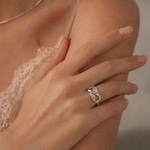 May include: A silver ring with a pink gemstone and wing-like designs. The ring is on a finger, and the background is a soft, neutral color. The ring's design is intricate and detailed.