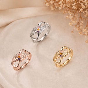 May include: Three rings with an infinity symbol design, each featuring a pair of wings and small gemstones. One ring is silver, another is rose gold, and the third is gold. The gemstones are pink, yellow, and purple.