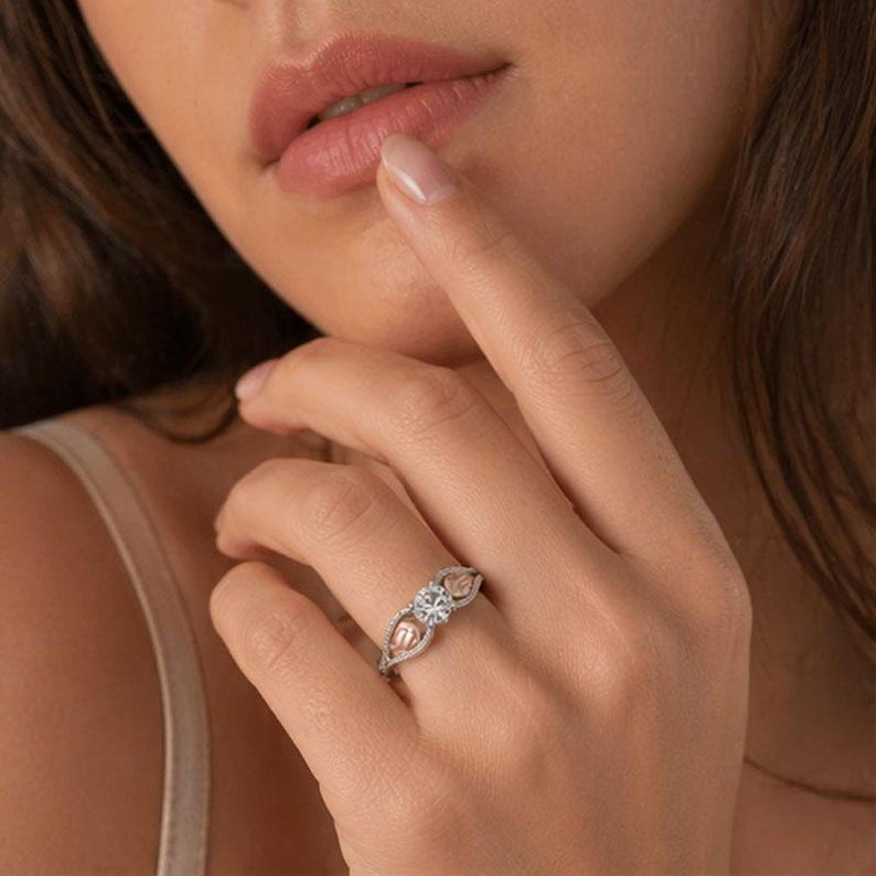 Puede incluir: Un anillo de plata con una gema central redonda y transparente, flanqueada por dos detalles en forma de coraz&oacute;n en oro rosa. La banda est&aacute; detallada con peque&ntilde;as piedras brillantes. El anillo est&aacute; en un dedo.