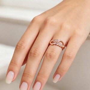 May include: Rose gold ring featuring an infinity symbol design with leaf accents. The ring has a small, colorful gemstone cluster at the center. The ring is displayed on a hand with manicured nails.