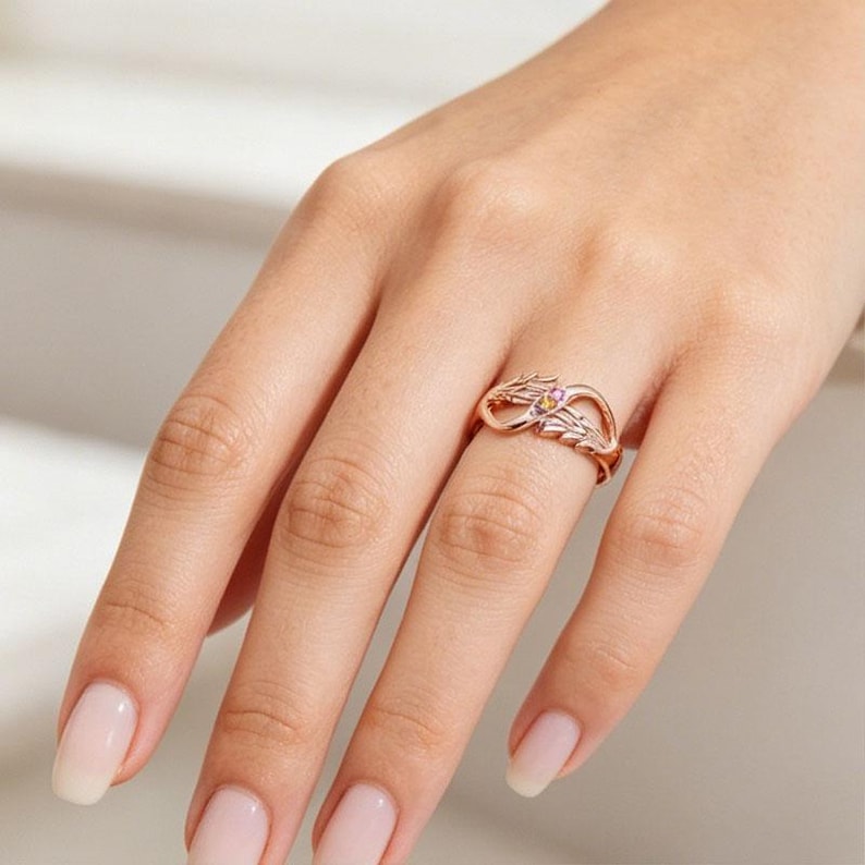 May include: Rose gold ring featuring an infinity symbol design with leaf accents. The ring has a small, colorful gemstone cluster at the center. The ring is displayed on a hand with manicured nails.