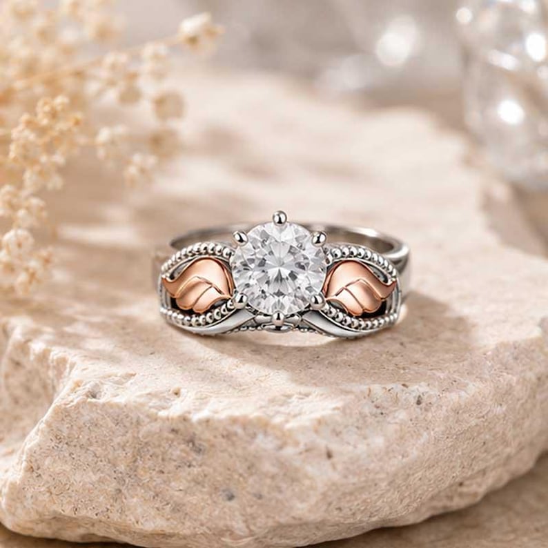 Puede incluir: Un anillo plateado con una gema redonda y transparente de gran tama&ntilde;o como pieza central. El anillo presenta detalles de alas en color oro rosa a ambos lados de la gema. El anillo est&aacute; sobre una superficie de piedra clara.