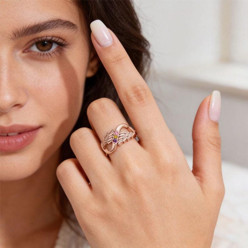 May include: Rose gold ring with an infinity symbol design, featuring small colorful gemstones. The ring has an open design with detailed wing-like elements on each side, and is worn on a finger.