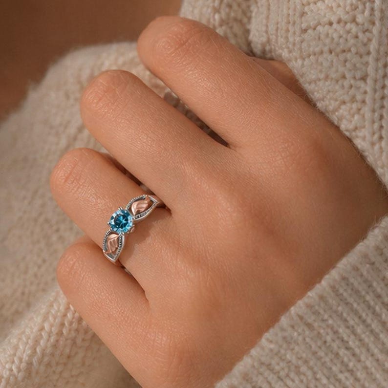 Puede incluir: Un anillo de plata con una piedra preciosa azul brillante y redonda en el centro. El anillo presenta un dise&ntilde;o en forma de hoja a cada lado de la piedra preciosa, con un acabado bicolor plateado y oro rosa. El anillo se lleva en un dedo.