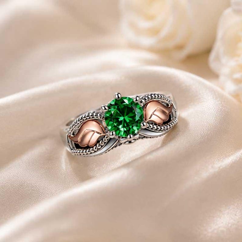 Puede incluir: Anillo de plata con una gran gema redonda de color verde esmeralda. El anillo presenta dos acentos de hojas de oro rosa a cada lado de la gema. La banda tiene un dise&ntilde;o detallado y con cuentas. El anillo est&aacute; sobre un fondo suave color crema.