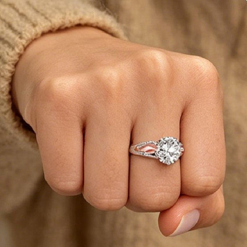 Puede incluir: Un anillo plateado con una gema grande, redonda y transparente. El anillo tiene un dise&ntilde;o de ca&ntilde;a dividida con un acento de color rosado. El anillo est&aacute; en un dedo.