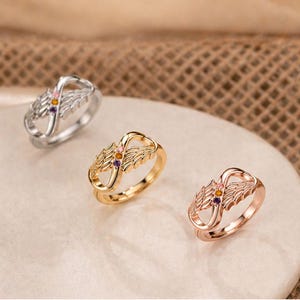 May include: Three rings with an infinity symbol design and wing accents. The rings are in silver, gold, and rose gold. Each ring has small, colorful gemstones. The rings are displayed on a light-colored surface.