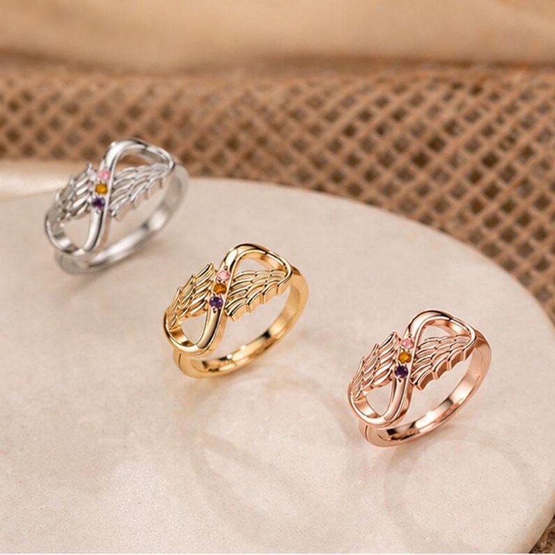 May include: Three rings with an infinity symbol design and wing accents. The rings are in silver, gold, and rose gold. Each ring has small, colorful gemstones. The rings are displayed on a light-colored surface.