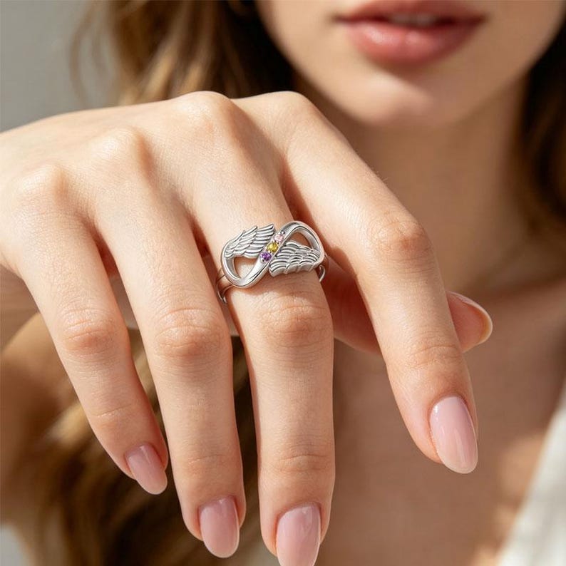 May include: A silver ring featuring an infinity symbol design with two sets of wings. The ring is adorned with small, colorful gemstones. The ring is a piece of jewelry.
