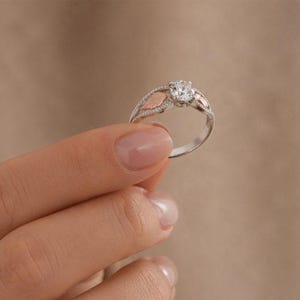 Puede incluir: Un anillo de plata con una gema redonda y transparente en el centro. El anillo presenta detalles en forma de hoja de oro rosa a ambos lados de la gema y peque&ntilde;as piedras transparentes a lo largo de la banda. El anillo es sostenido por una mano.