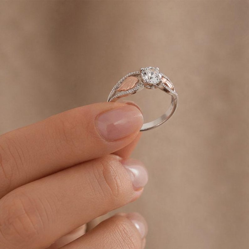 Puede incluir: Un anillo de plata con una gema redonda y transparente en el centro. El anillo presenta detalles en forma de hoja de oro rosa a ambos lados de la gema y peque&ntilde;as piedras transparentes a lo largo de la banda. El anillo es sostenido por una mano.