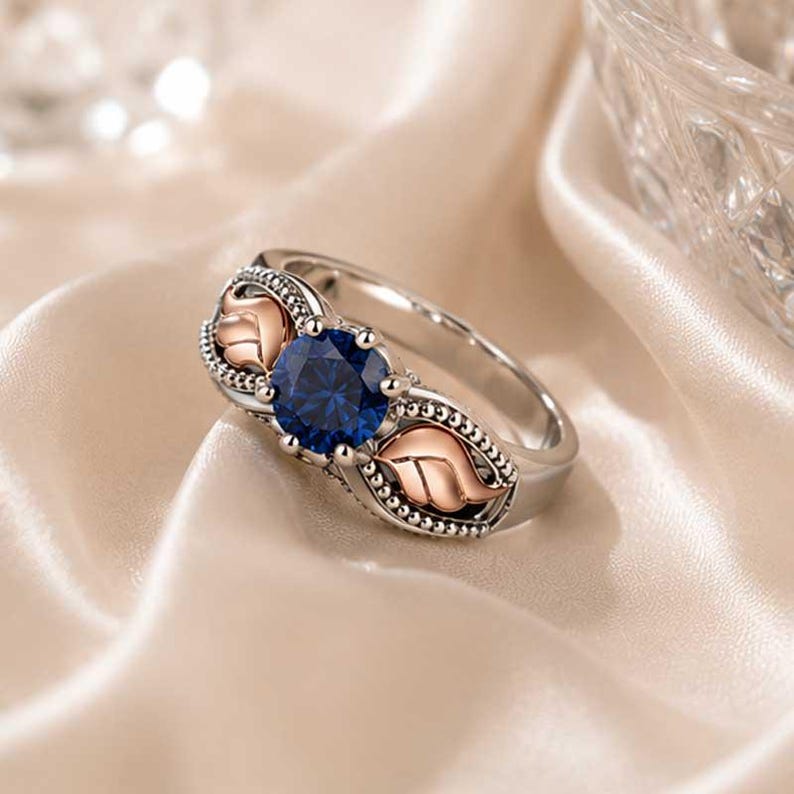 Puede incluir: Anillo plateado con una gema azul oscuro grande y redonda. El anillo presenta detalles de hojas de oro rosa a ambos lados de la gema. La banda es plateada con un dise&ntilde;o de cuentas.