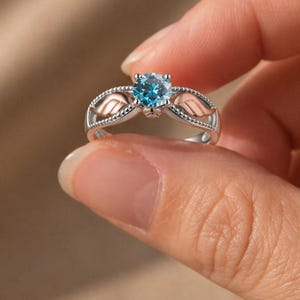 Puede incluir: Un anillo plateado con una piedra preciosa redonda de color azul claro en el centro. El anillo presenta un dise&ntilde;o de dos alas doradas a cada lado de la piedra preciosa. El anillo se sostiene entre los dedos.