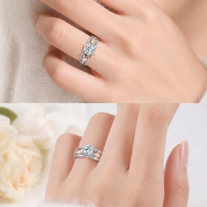 Puede incluir: Un anillo plateado con una piedra preciosa grande, redonda y transparente en el centro. El anillo tiene un dise&ntilde;o decorativo con detalles en oro rosa a ambos lados de la piedra central. El anillo se lleva en un dedo.