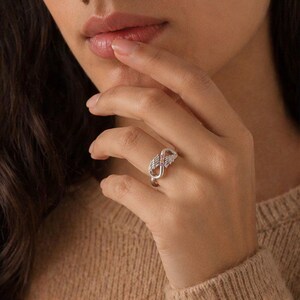 May include: A silver ring featuring a double infinity symbol design with small pink gemstones. The ring is worn on a finger, showcasing its intricate details and elegant style. The ring's design incorporates a delicate, openwork pattern.