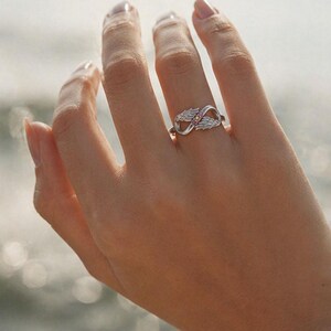 May include: A silver ring featuring an infinity symbol design with wing accents and a small pink gemstone. The ring is worn on a finger, with a blurred background of water.