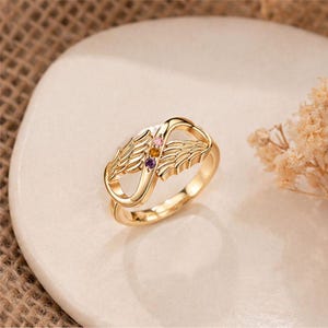 May include: A gold-colored ring with an infinity symbol design, featuring wing details on each side. The ring is adorned with three small gemstones in pink, yellow, and purple. The ring is displayed on a white surface.