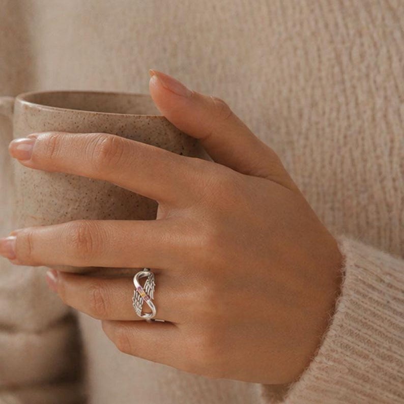 May include: A silver ring with an infinity symbol design, featuring small pink stones. The ring is worn on a finger, with a beige sweater and a ceramic mug in the background.
