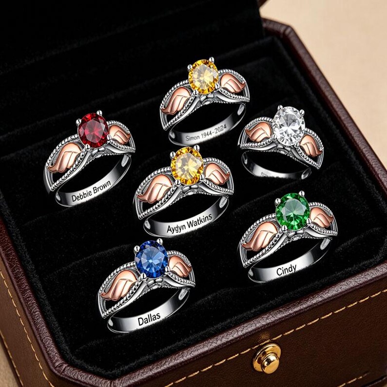 Puede incluir: Una colecci&oacute;n de anillos personalizados con piedras preciosas de varios colores, incluyendo rojo, amarillo, blanco, verde y azul. Cada anillo tiene una banda plateada con alas de oro rosa y nombres o fechas grabadas. Los anillos se exhiben en una caja de joyer&iacute;a marr&oacute;n.