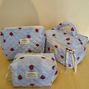 May include: Three blue and white patterned cosmetic bags with a strawberry design. The bags are quilted and have a zipper closure. The text "milobliss find your fire" is on a small label on each bag.