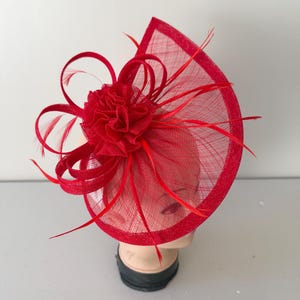 May include: A vibrant red fascinator, featuring a semi-circular mesh base with a structured frame. The headpiece is adorned with a central floral embellishment and delicate feather accents, creating a dramatic and elegant look.