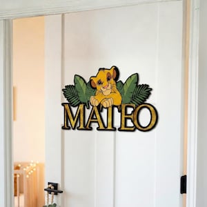 May include: A wooden door sign featuring a cartoon lion cub with the name "MATEO" in gold lettering. The lion is yellow and brown, set against green leaves. The sign is mounted on a white door.