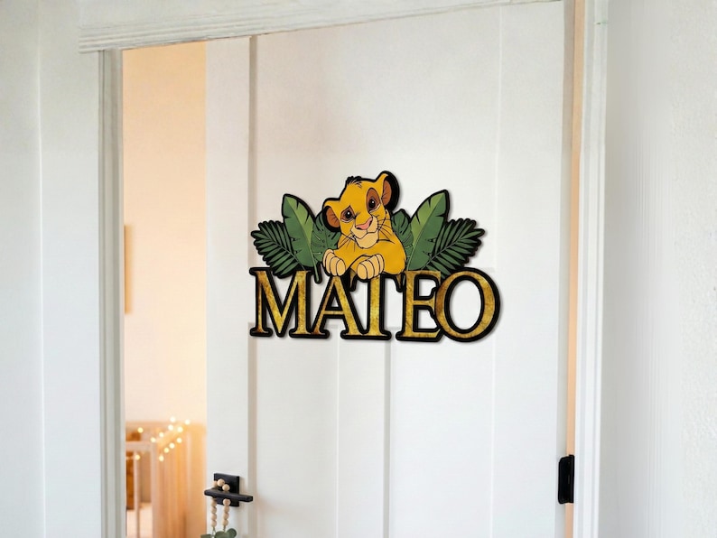 May include: A wooden door sign featuring a cartoon lion cub with the name "MATEO" in gold lettering. The lion is yellow and brown, set against green leaves. The sign is mounted on a white door.