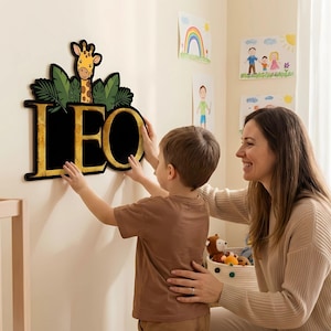 May include: A wooden wall decoration spelling out the name "LEO" in gold lettering. The letters are set against a black background, with a giraffe and green leaves above. A child and an adult are shown interacting with the decoration.