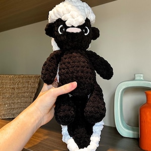 Peut inclure: Une peluche de mouffette faite à la main au crochet. La peluche a un corps noir avec une rayure blanche et une tête blanche et duveteuse. Elle a de petites oreilles noires, un nez rose et des yeux blancs. La mouffette est tenue dans une main.